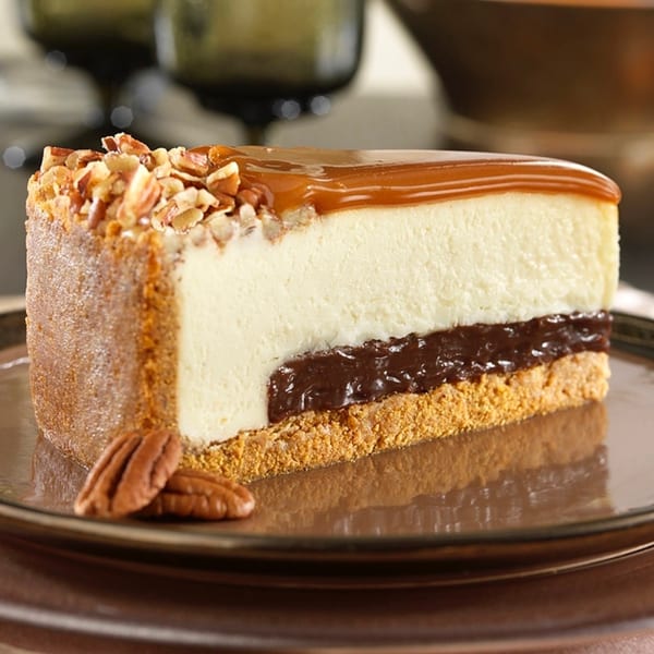 Slice of cheesecake with caramel, chopped pecans, and a chocolate layer on a brown plate.